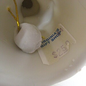 May include: Close-up of a white ceramic object with a small, attached white ornament and a gold string. A price tag with the text "VIRGINIA'S GIFT SHOP" is visible. The object appears to be a decorative item.