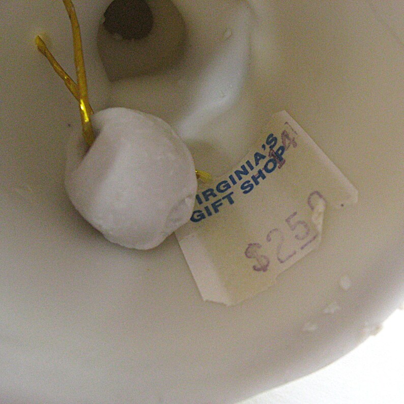 May include: Close-up of a white ceramic object with a small, attached white ornament and a gold string. A price tag with the text "VIRGINIA'S GIFT SHOP" is visible. The object appears to be a decorative item.