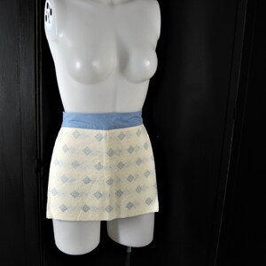 May include: A white apron with a blue waistband and a light blue geometric pattern. The apron is on a white mannequin.