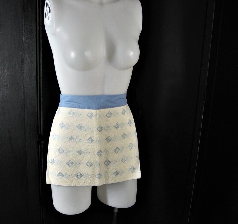 May include: A white apron with a blue waistband and a light blue geometric pattern. The apron is on a white mannequin.