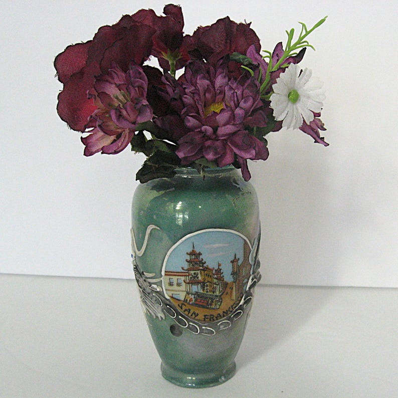 Pode incluir: Um vaso verde com um design de S&atilde;o Francisco, segurando um buqu&ecirc; de flores roxas escuras e brancas. O vaso tem um design prateado met&aacute;lico ao redor da imagem de S&atilde;o Francisco e ao redor do pr&oacute;prio vaso.