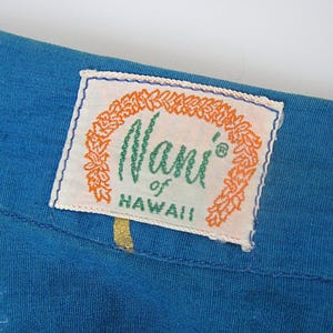 May include: A close-up shot of blue fabric with a rectangular label. The label reads "Nani of Hawaii" in green script, framed by orange decorative elements on a white background. The label is edged with blue stitching.