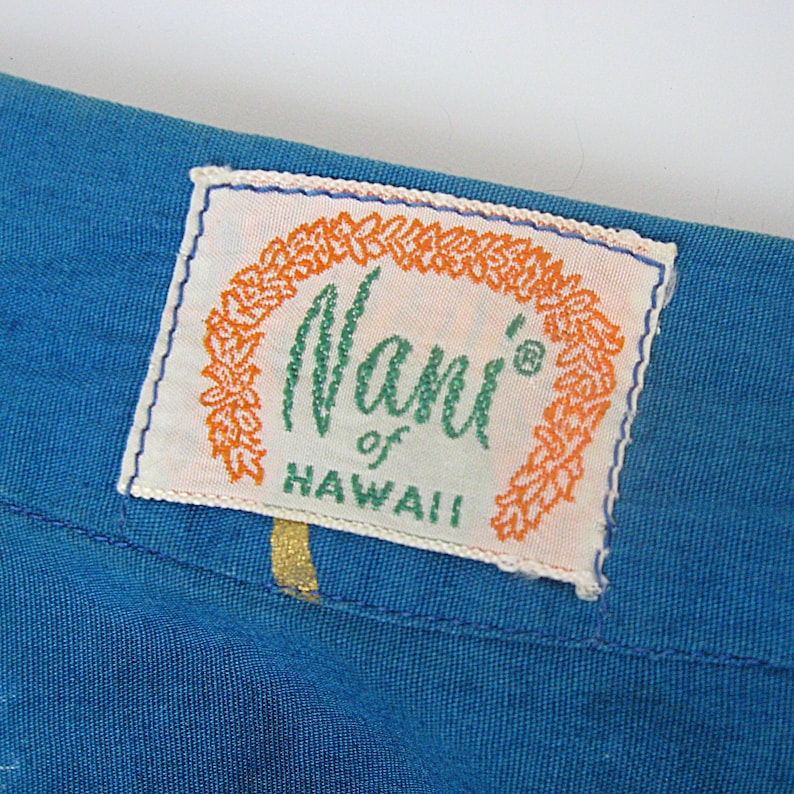 May include: A close-up shot of blue fabric with a rectangular label. The label reads "Nani of Hawaii" in green script, framed by orange decorative elements on a white background. The label is edged with blue stitching.