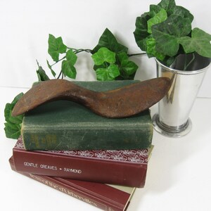 May include: A rusty metal shoe stretcher, shaped like a curved bar, resting on top of a stack of three books. The books are green, burgundy, and brown. The top book has the title "Gentle Greaves - Raymond" printed on the spine. The shoe stretcher is a vintage tool used to stretch shoes.