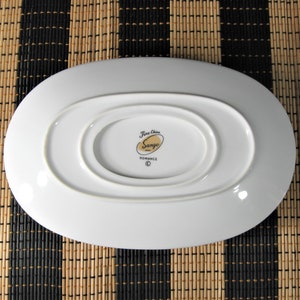 K&ouml;nnte beinhalten: Wei&szlig;e ovale Servierplatte mit einem erh&ouml;hten Mittelteil. Die Unterseite der Platte tr&auml;gt ein goldenes Logo mit der Aufschrift "Fine China Sango Romance" mit einem Copyright-Symbol.