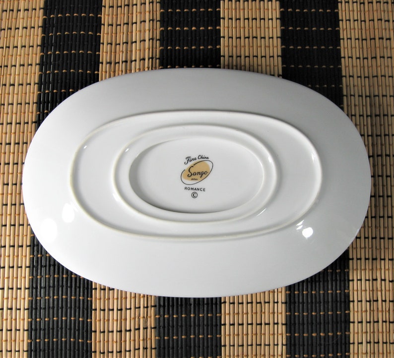 K&ouml;nnte beinhalten: Wei&szlig;e ovale Servierplatte mit einem erh&ouml;hten Mittelteil. Die Unterseite der Platte tr&auml;gt ein goldenes Logo mit der Aufschrift "Fine China Sango Romance" mit einem Copyright-Symbol.