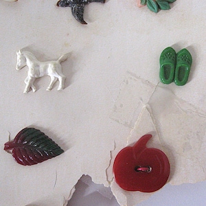 May include: A collection of assorted decorative buttons featuring a white horse, a pair of green clogs, a dark red leaf, and a bright red apple-shaped button. These small, molded plastic items are arranged on a light-colored, textured surface.