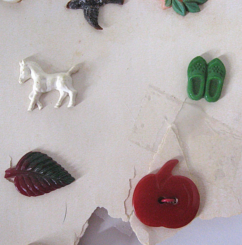 May include: A collection of assorted decorative buttons featuring a white horse, a pair of green clogs, a dark red leaf, and a bright red apple-shaped button. These small, molded plastic items are arranged on a light-colored, textured surface.