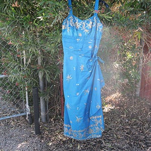 May include: A blue dress with a wrap-style skirt and spaghetti straps. The dress has a gold floral pattern and is hanging outside on a hanger. The dress is made of a lightweight fabric.