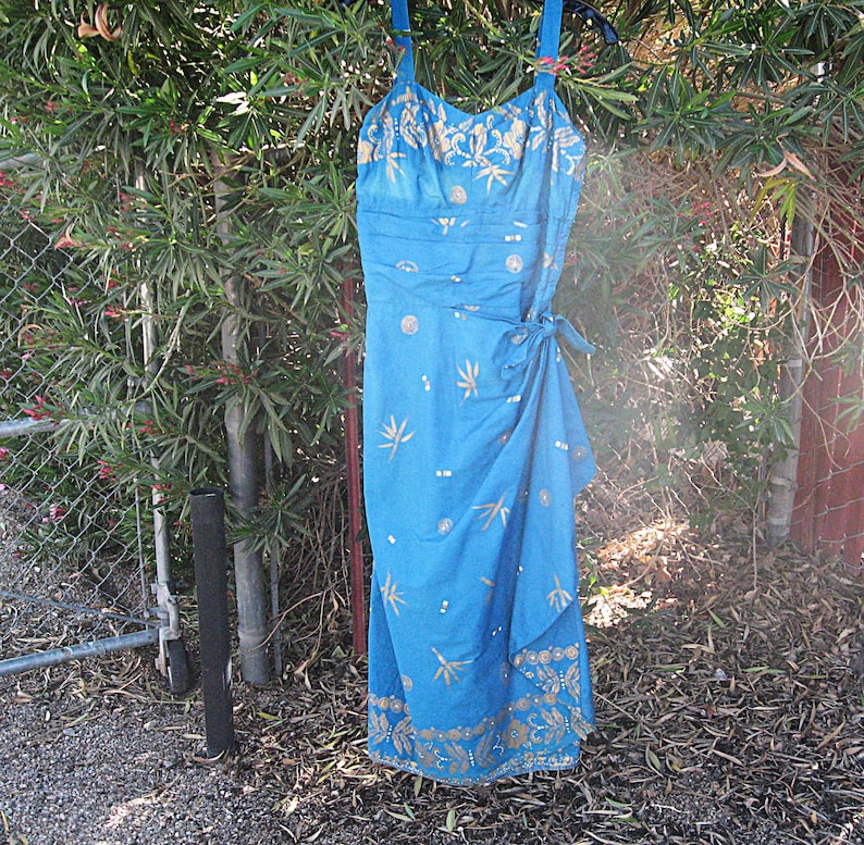 May include: A blue dress with a wrap-style skirt and spaghetti straps. The dress has a gold floral pattern and is hanging outside on a hanger. The dress is made of a lightweight fabric.
