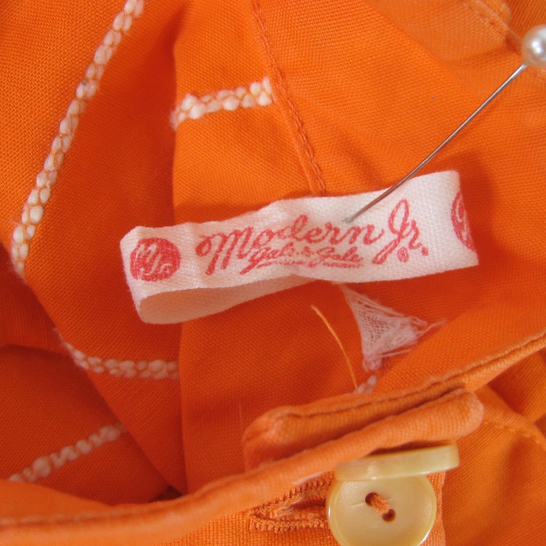 May include: Close-up of a white label with red text that reads "Modern Jr. Girls & Boys" on an orange fabric background. The label is attached to a garment with a white thread.