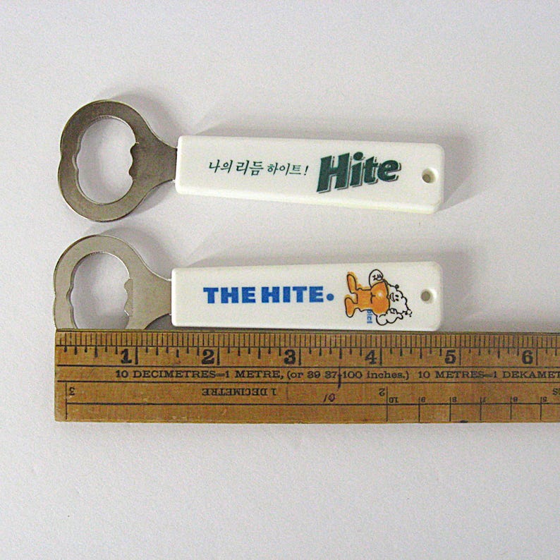 May include: Two white bottle openers with metal ends. One has the word "Hite" in green and Korean text. The other says "THE HITE." with a cartoon character. A wooden ruler is below the openers.