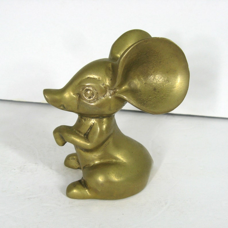 May include: A small, gold-colored brass mouse figurine with large ears. The mouse is sitting on its hind legs with its front paws raised.