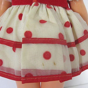 May include: A doll skirt made of sheer off-white fabric featuring a red polka dot pattern. The skirt has a gathered waist with a solid red fabric band and two horizontal red ribbon stripes running across the lower portion of the garment.