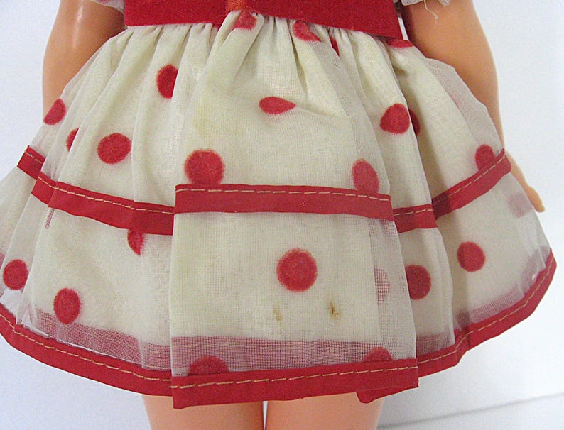 May include: A doll skirt made of sheer off-white fabric featuring a red polka dot pattern. The skirt has a gathered waist with a solid red fabric band and two horizontal red ribbon stripes running across the lower portion of the garment.