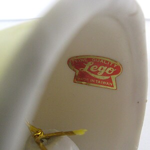 May include: Close-up of a cream-colored ceramic object with a red and gold sticker that reads "Fine Quality Lego Made in Taiwan". The object has a smooth, curved surface.