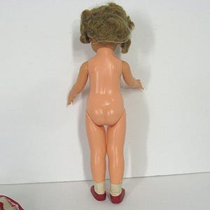 May include: A plastic doll viewed from the back, featuring light skin tone, curly brown hair, and wearing white socks with dark red shoes. The doll has articulated arms and legs, standing upright against a plain, neutral white background.