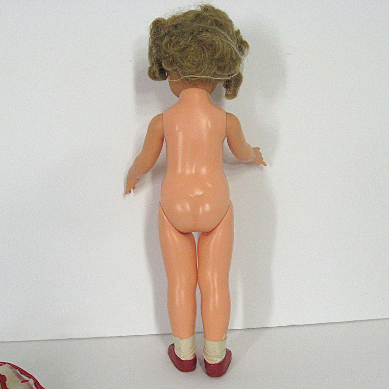 May include: A plastic doll viewed from the back, featuring light skin tone, curly brown hair, and wearing white socks with dark red shoes. The doll has articulated arms and legs, standing upright against a plain, neutral white background.