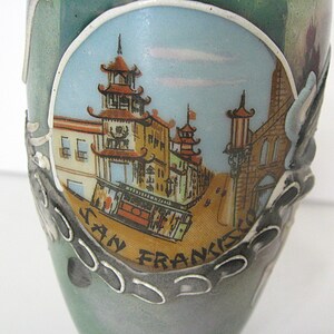 Pode incluir: Um vaso de cer&acirc;mica com uma cena pintada de Chinatown em S&atilde;o Francisco. A imagem apresenta edif&iacute;cios com telhados em estilo pagode, uma rua com um telef&eacute;rico e as palavras "SAN FRANCISCO". O vaso tem um esquema de cores verde-azulado e cinza.