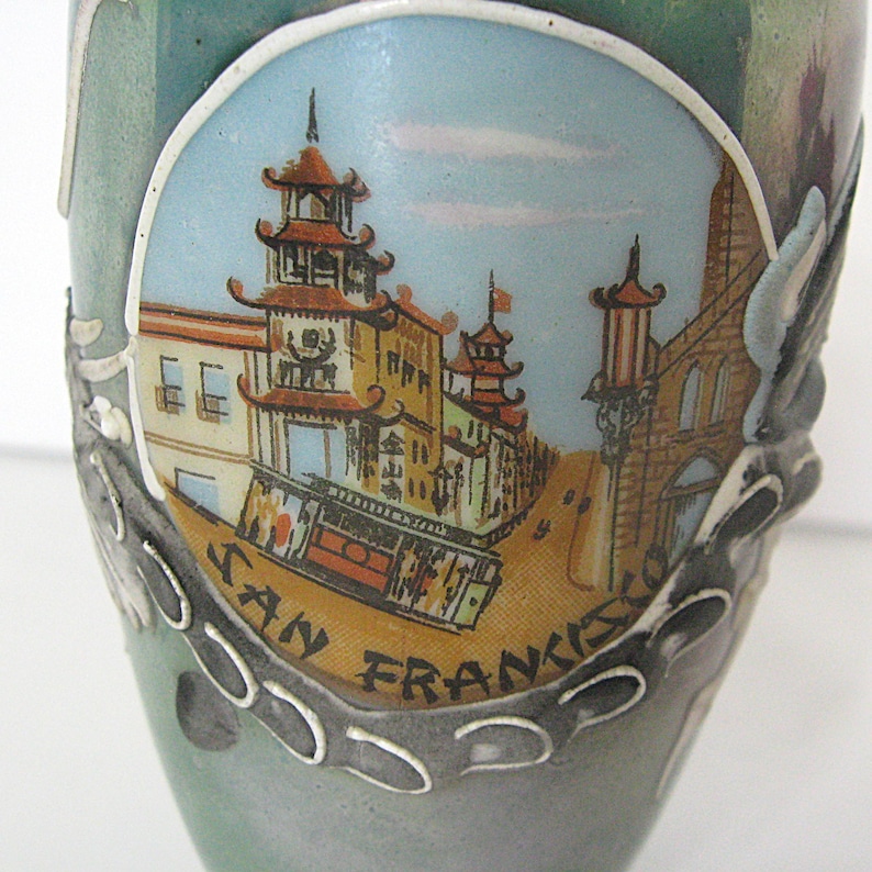 Pode incluir: Um vaso de cer&acirc;mica com uma cena pintada de Chinatown em S&atilde;o Francisco. A imagem apresenta edif&iacute;cios com telhados em estilo pagode, uma rua com um telef&eacute;rico e as palavras "SAN FRANCISCO". O vaso tem um esquema de cores verde-azulado e cinza.