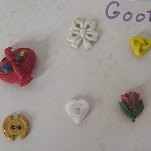 May include: A collection of six decorative buttons including a red basket, a white bow, a yellow knot, a gold circular button, a white heart-shaped button, and a red tulip with green leaves. The word Goofi is partially visible in the top right corner.