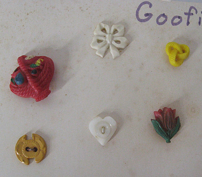 May include: A collection of six decorative buttons including a red basket, a white bow, a yellow knot, a gold circular button, a white heart-shaped button, and a red tulip with green leaves. The word Goofi is partially visible in the top right corner.