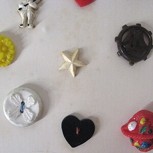 May include: A collection of assorted buttons including a yellow floral button, a white star, a dark brown ship wheel, a white button with a four-leaf design, a black heart-shaped button, a red basket-shaped button, and a small white figure.