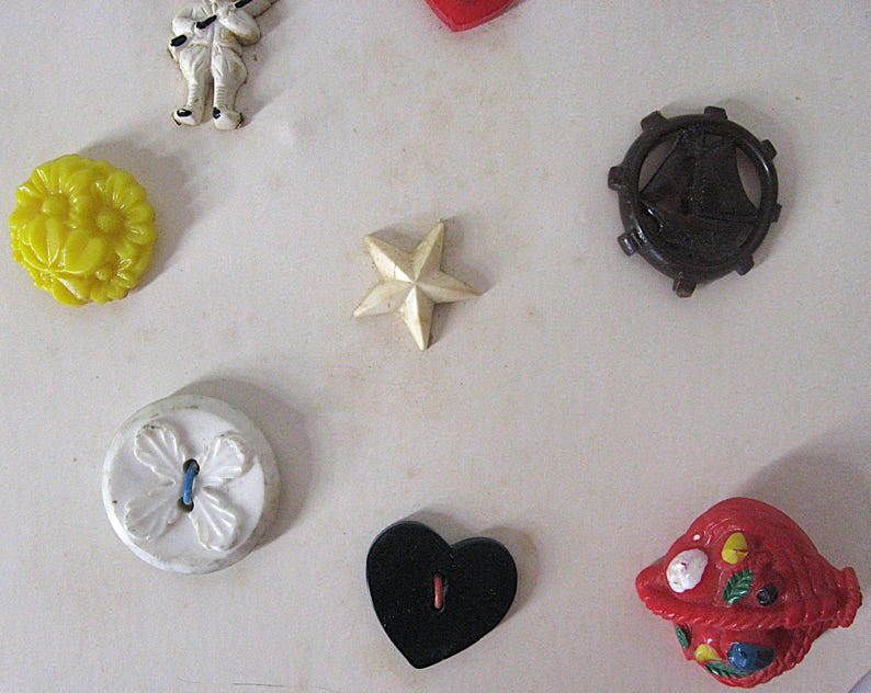 May include: A collection of assorted buttons including a yellow floral button, a white star, a dark brown ship wheel, a white button with a four-leaf design, a black heart-shaped button, a red basket-shaped button, and a small white figure.