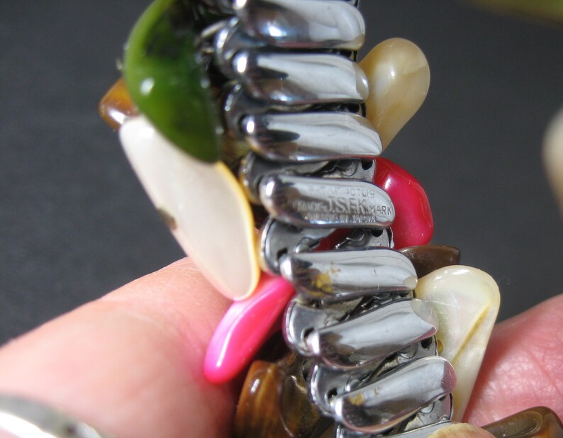 May include: A silver expandable bracelet with a variety of colorful stones, including green, white, pink, and brown. The bracelet is marked "J.S.K. MARK" and "PAT. PENDING".