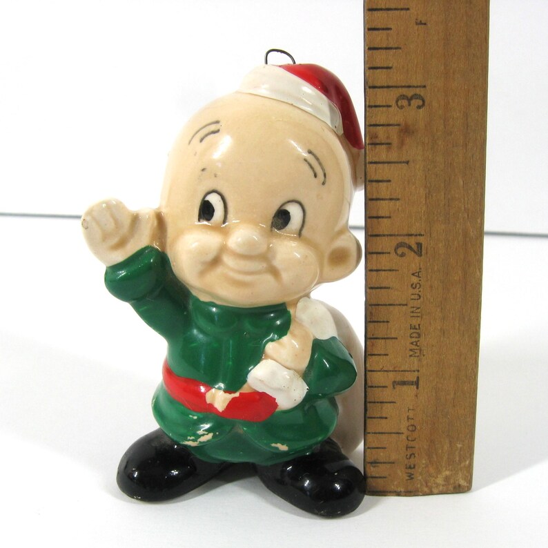 May include: A ceramic Christmas ornament depicting a cartoon character wearing a green and red outfit and a Santa hat. The character is waving with one hand and holding a bag in the other. The ornament is approximately 5 cm tall.