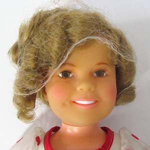 May include: A doll head with light brown curly hair styled with a fine white netting veil. The doll features brown eyes, rosy cheeks, and a smiling mouth with visible white teeth. The doll wears a white garment with red accents around the neck.