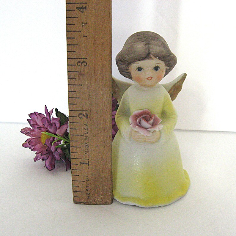 May include: A ceramic angel figurine, holding a pink rose, with a yellow dress and brown hair. The figurine is approximately 3 inches tall, measured with a wooden ruler. A cluster of purple flowers is to the left.