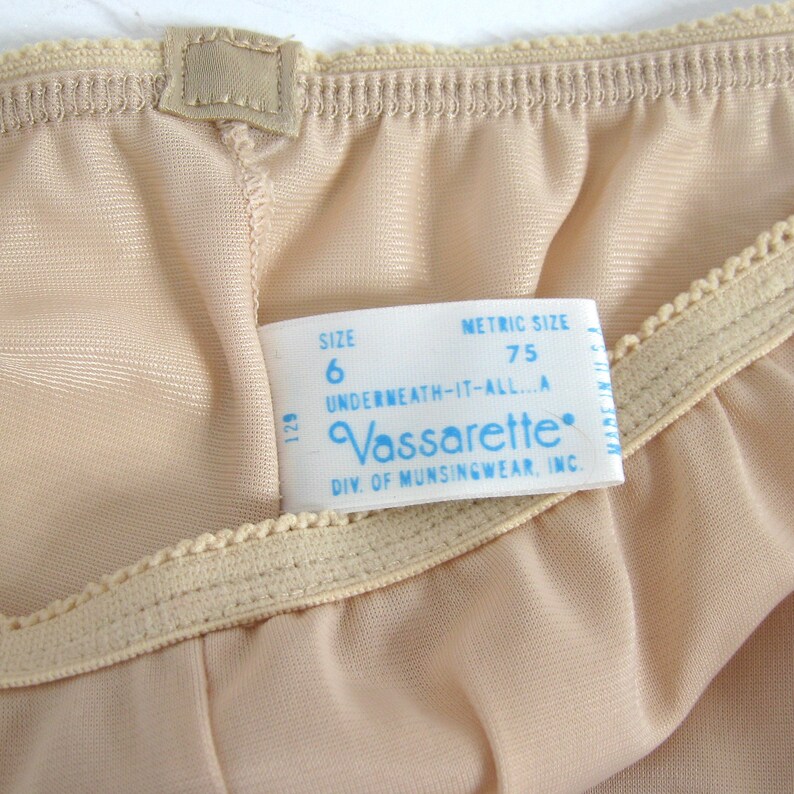 May include: A beige underwear label with blue text that reads "Vassarette" and "Underneath-It-All...A" with the size "6" and metric size "75".