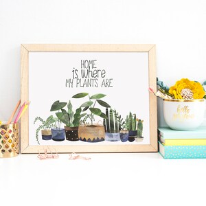 以下が含まれることがあります： 「Home is where my plants are」というテキストと、鉢植えの植物の水彩画が描かれた額装されたプリント。プリントは、金の鉛筆立て、2つの金のクリップ、そして黄色い花と金の「hello sunshine」という文字が書かれた白いマグカップが置かれた白い机の上に置かれています。