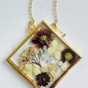 May include: A diamond-shaped glass frame with a thin gold metal border, suspended by a gold chain. Inside the frame are pressed dried flowers, including dark red daisy-like blooms and pale cream-colored hydrangea petals arranged on a white background.