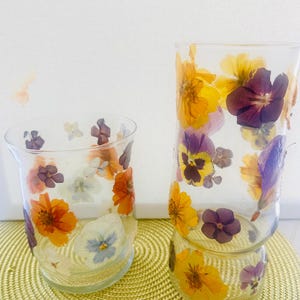 May include: Two clear glass tumblers decorated with pressed, dried flowers in shades of yellow, orange, purple, and white. The flowers are embedded on the exterior surface of the glass, featuring pansies and other small blossoms in a scattered pattern.