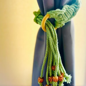 May include: A green rope curtain tieback with a wooden ring and wooden beads. The tieback is attached to a blue curtain.