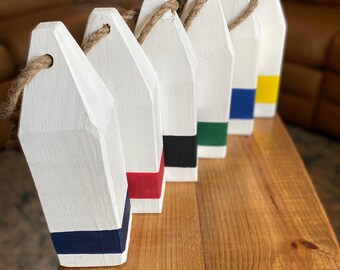 May include: A row of white wooden buoys with colorful bands of navy, red, black, green, blue, and yellow. Each buoy has a rope attached for hanging. The buoys are arranged on a wooden surface.