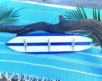 May include: A surfboard-shaped wall-mounted coat rack. The rack is painted white with blue stripes and has three silver metal hooks. A piece of driftwood and palm fronds are in the background.