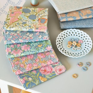 May include: A collection of patterned fabric squares in various floral and botanical designs, including pink peonies and blue flowers. A spool of thread and decorative buttons are also present, arranged on a light blue surface.