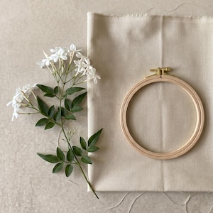 Peut inclure: Un tambour à broder rond en bois avec une vis de tension en laiton posé sur un morceau de tissu rectangulaire beige, accompagné d'une branche de fleurs de jasmin blanches avec des feuilles vertes placée à gauche du tissu et du tambour.
