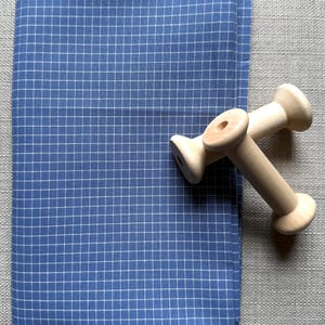 May include: A folded blue fabric with a white grid pattern, next to two wooden spools. The fabric is a solid blue with a white grid overlay. The spools are light-colored wood, with a cylindrical body and flared ends.