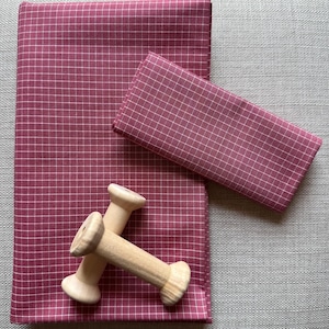 May include: A flat lay featuring a burgundy fabric with a white grid pattern, a smaller matching folded fabric, and two wooden spools. The fabrics are arranged on a neutral gray surface, creating a visually appealing composition.