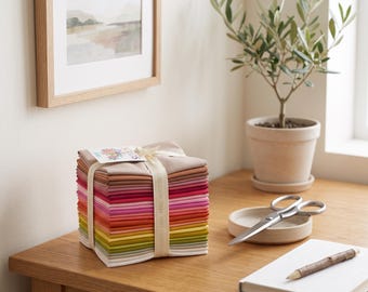 Op de afbeelding: Een stapel gevouwen vierkante stoffen in een kleurverloop van wit en beige tot roze, rood, oranje, geel en groen. Het stoffen pakket is samengebonden met een crèmekleurig lint en heeft een klein rechthoekig label aan de bovenzijde.