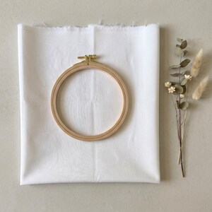 Puede incluir: Un bastidor de bordado circular de madera con un tornillo de tensión de latón centrado sobre una pieza rectangular de tela blanca. Junto a la tela hay un pequeño y delicado arreglo de hojas de eucalipto secas y hierba de cola de conejo.