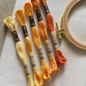 May include: A collection of embroidery floss in shades of yellow and orange, with a wooden embroidery hoop and a piece of white fabric. The floss is labeled with numbers and brand information. Ideal for embroidery and cross-stitch projects.