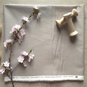 May include: A neutral-toned fabric from the Tilda Quilt Collection, with a sprig of pink cherry blossoms and two wooden spools. The fabric has a textured appearance and a white border with text. The fabric is labeled as "Mouse".