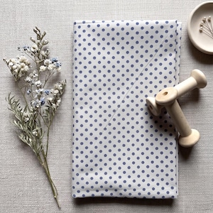 May include: A folded white fabric with a blue polka dot pattern. A sprig of dried flowers and a small wooden spool are next to the fabric. A small, off-white dish with tiny white flowers is also in the image.