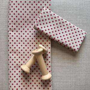 May include: A folded fabric and a stack of fabric with a red polka dot pattern on a cream background. Two wooden spools are in the foreground. The fabric is likely for sewing or crafting projects.