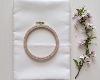 May include: A folded white fabric piece featuring a horizontal frayed edge and a thin pink stripe, topped with a circular wooden embroidery hoop with a brass tension screw, accompanied by a flowering branch with light pink blossoms.
